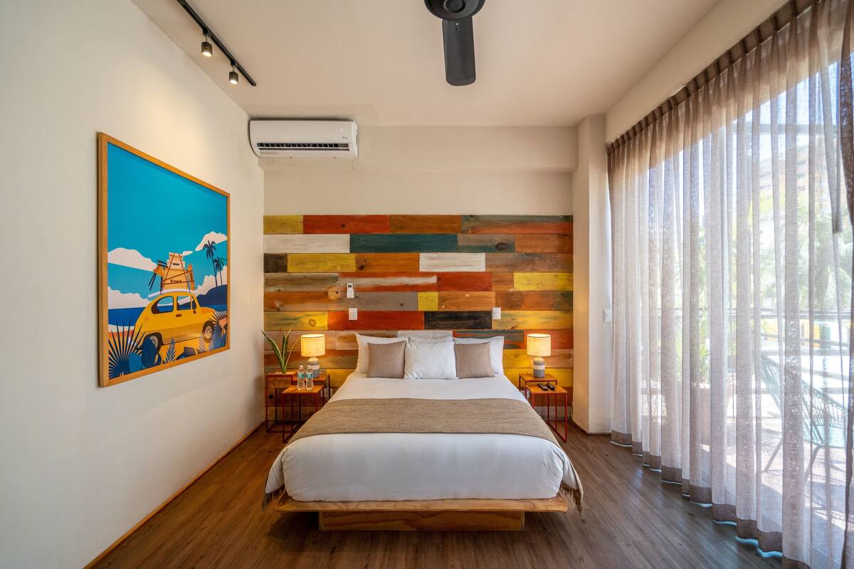 A modern hotel room with a wooden bed, colorful wooden wall paneling, a painting on the wall, and a large window with curtains.