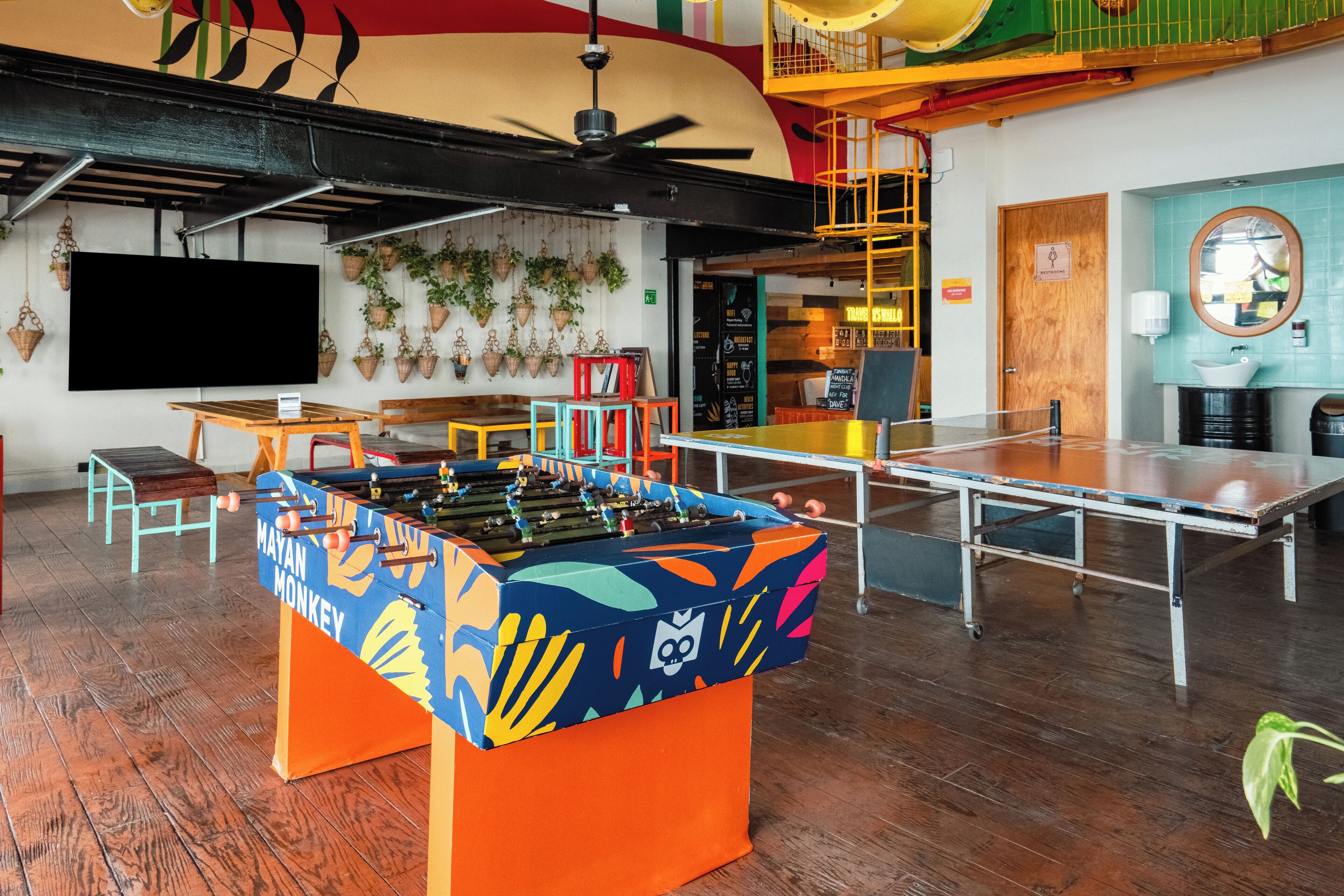 A room with a foosball table, ping pong table, and a TV screen.