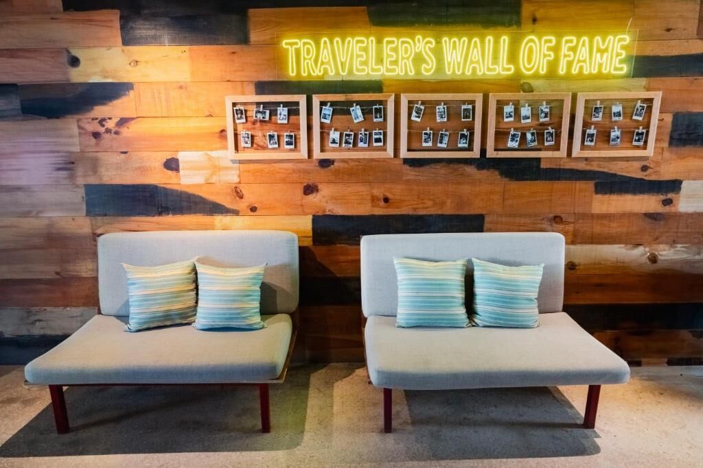 A room with two grey sofas, a neon sign reading "TRAVELER'S WALL OF FAME", and a wooden wall with framed photos.