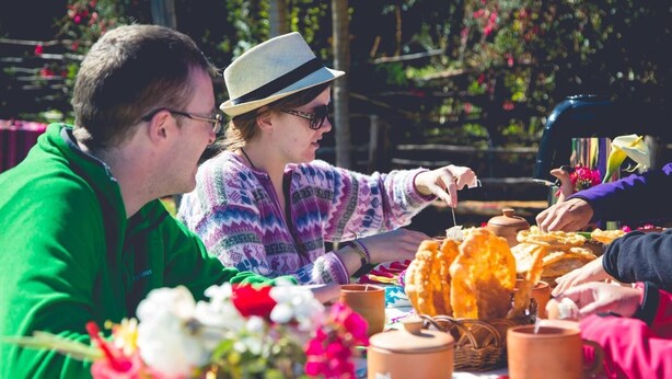 Een groep mensen zit aan een tafel met eten en drinken, omringd door bloemen en potten.