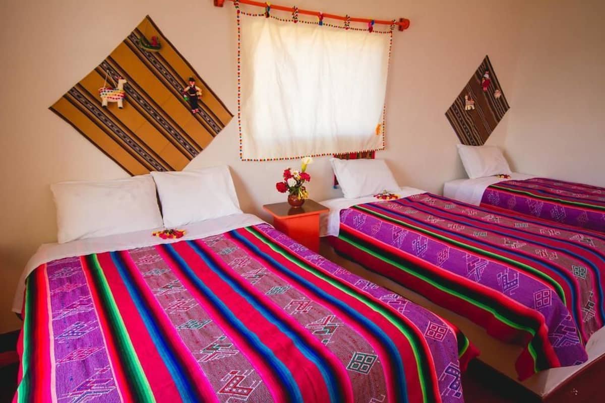 A hotel room with two beds, colorful patterned bedspreads, a small table with a vase of flowers, and decorative wall hangings.