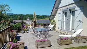 A patio with a table and chairs, surrounded by wooden planters with flowers, and a building with large windows and a door. - Chambres d'hôtes Anousta (Loubajac)