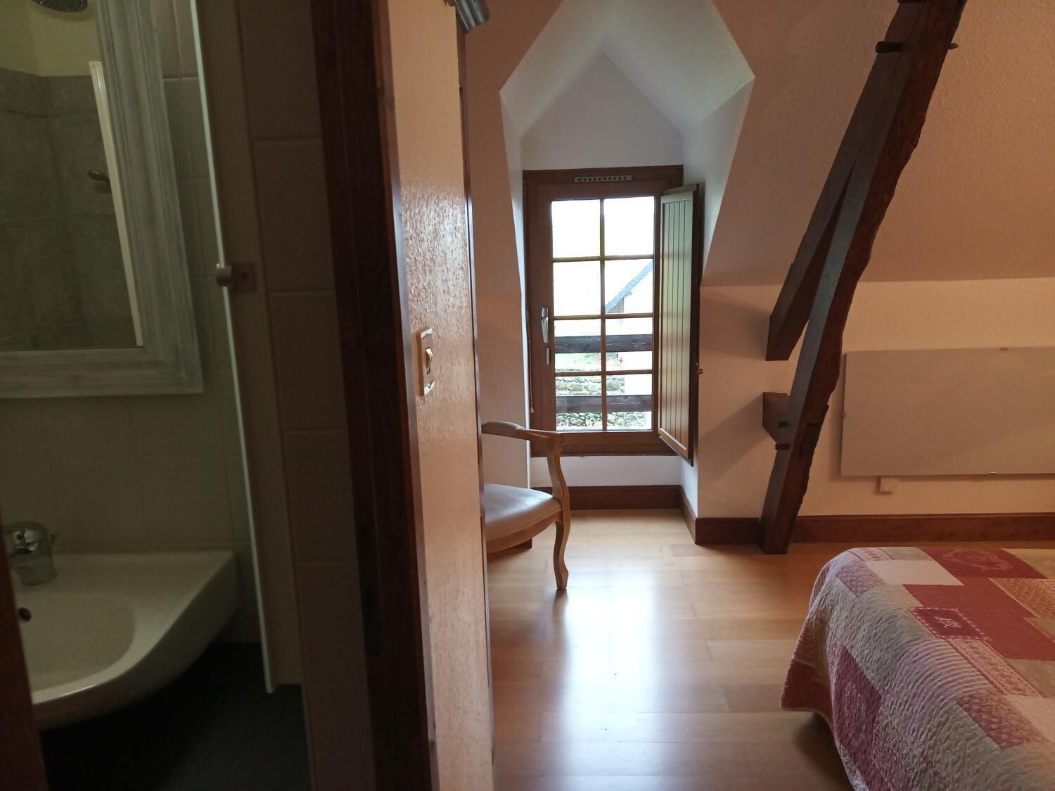 A bedroom with a wooden floor, a window with a view, a bed, and a door leading to another room.