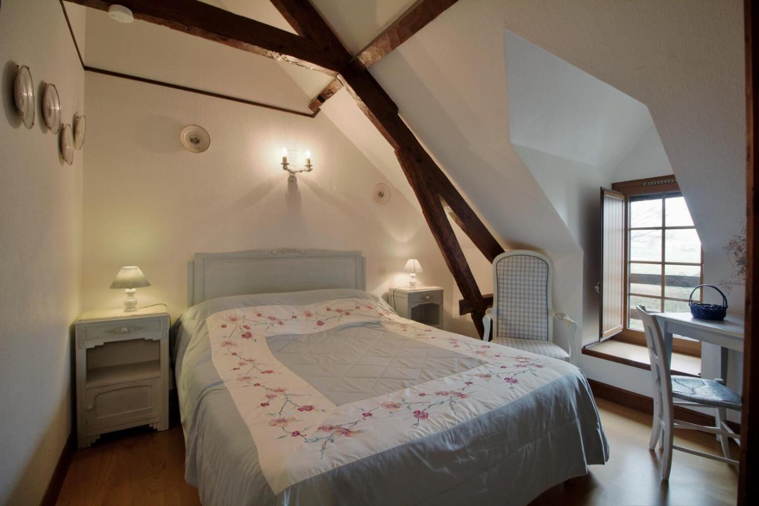Un dormitorio con techo inclinado, una cama con ropa de cama floral, lámparas de noche, una silla y un ventanal con vistas.