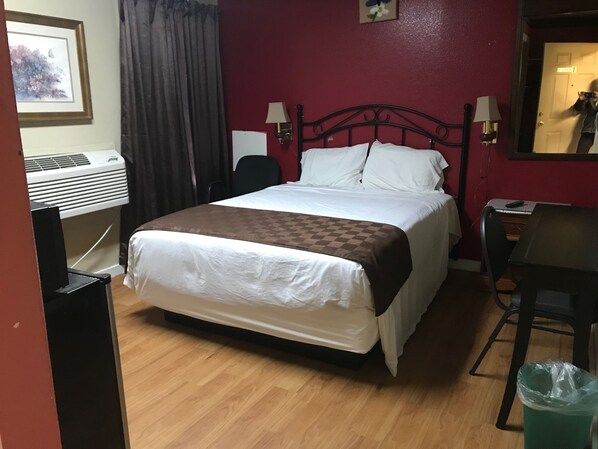 A hotel room with a bed, a desk, a chair, a trash bin, and a wall-mounted air conditioner.