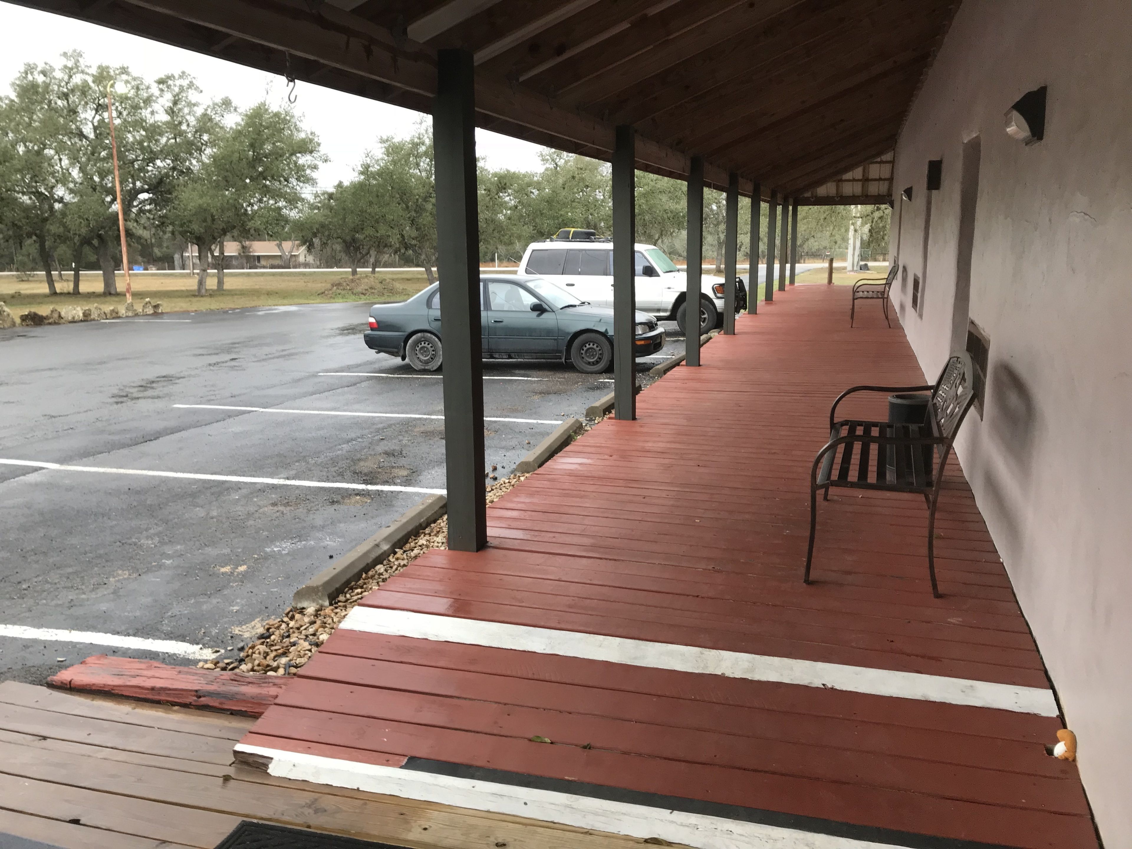 A covered outdoor area with a red wooden deck, a row of benches, and a parking lot with cars.