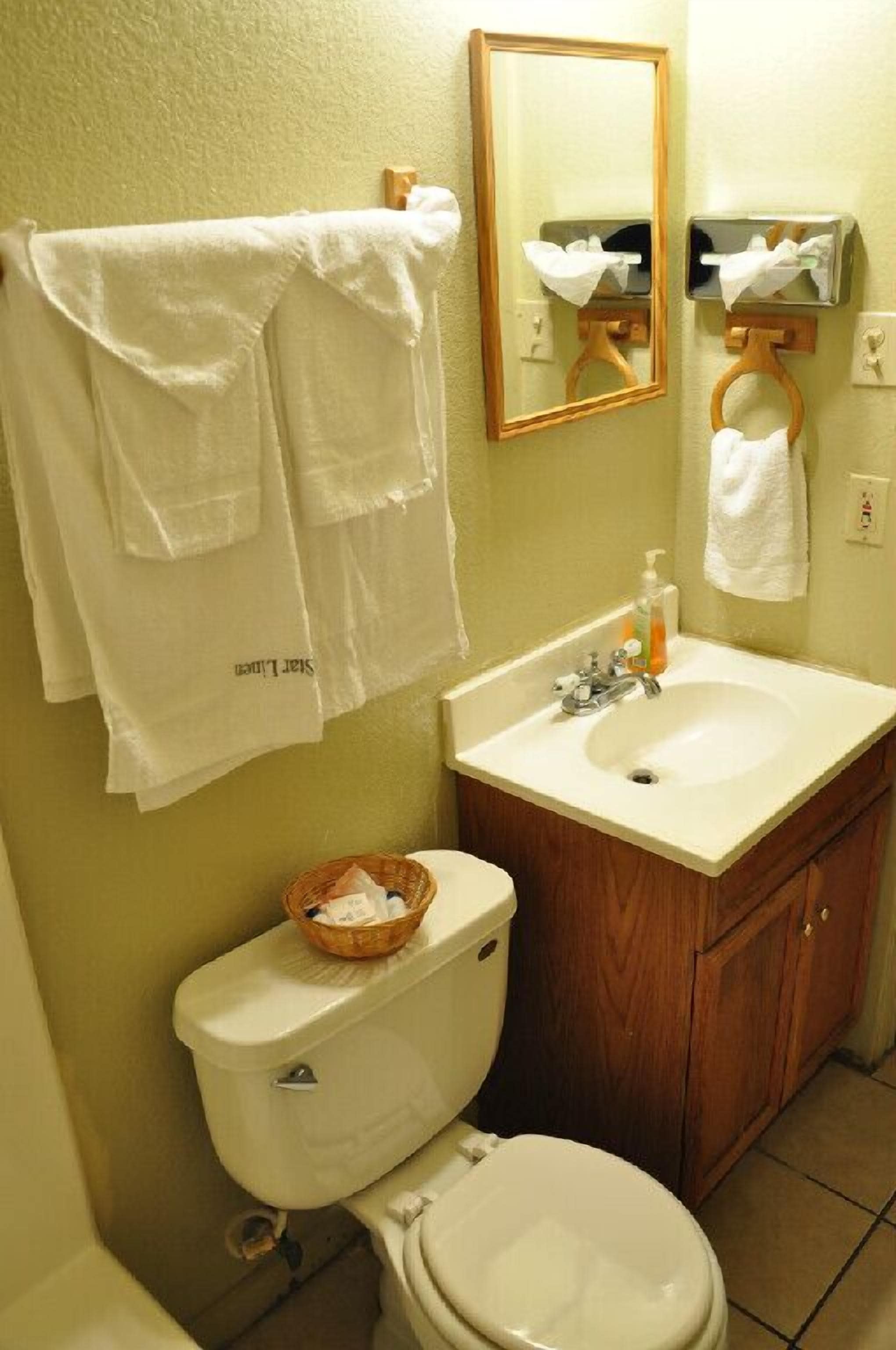 A bathroom with a toilet, sink, and mirror. There are towels hanging on the wall and a basket on the toilet.