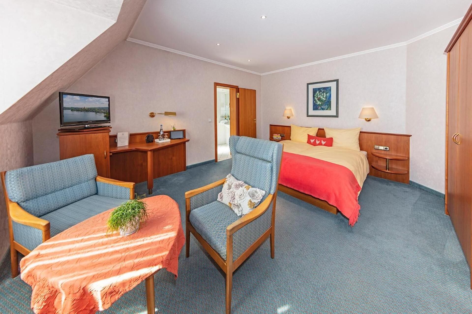 Quarto de hotel com uma cama, duas poltronas, uma mesinha com uma planta, uma escrivaninha com uma televisão e uma porta para o banheiro.