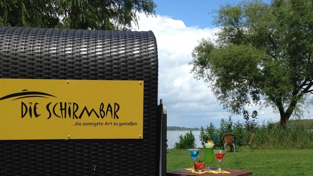 A covered outdoor seating area with a sign reading "Die Schirmbar" and a table with drinks. - Wittlers Hotel (Ratzeburg)