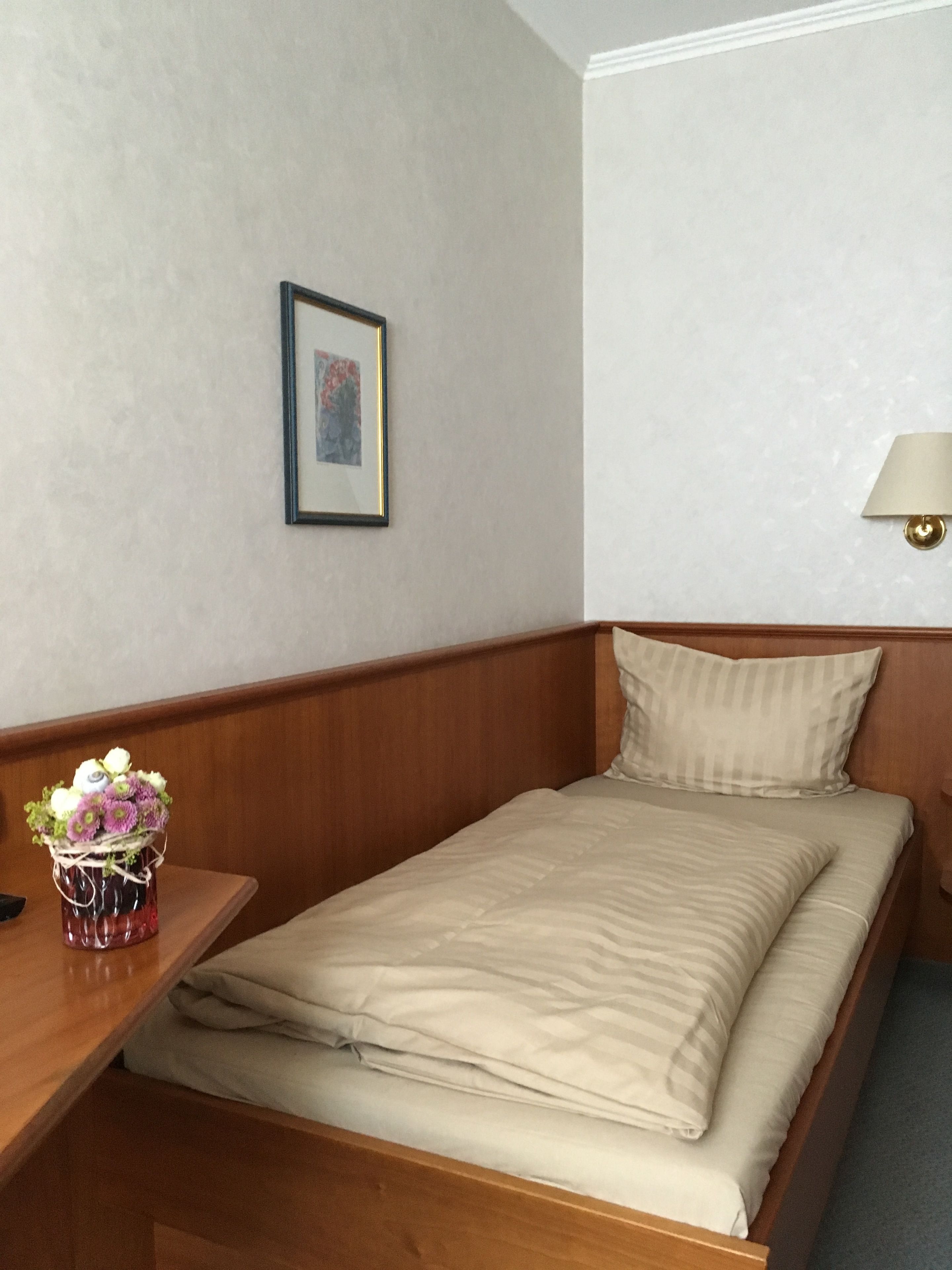 A single bed with a wooden headboard, a bedside table with a vase of flowers, a framed picture on the wall, and a wall-mounted lamp.