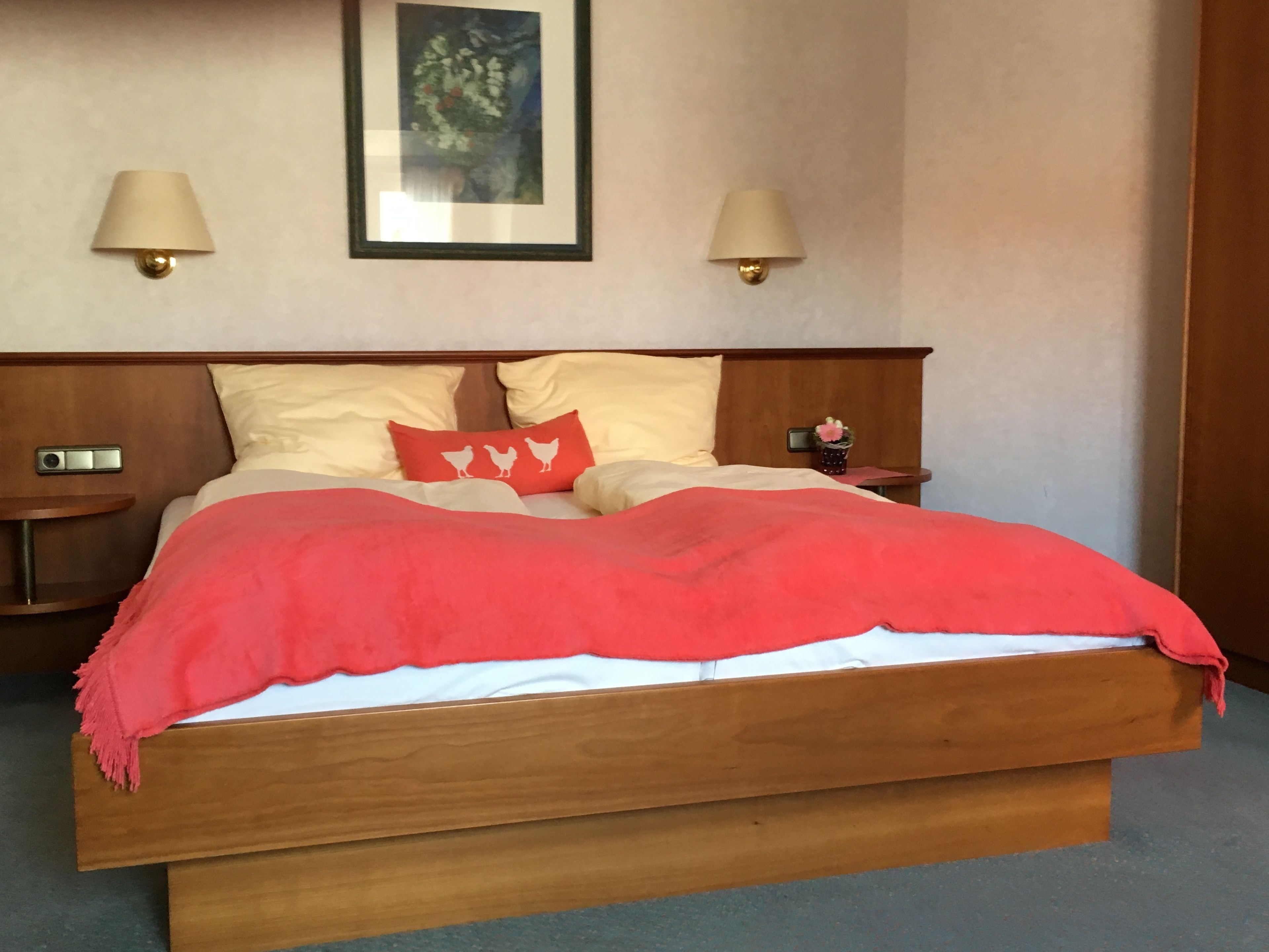 A hotel room with a wooden bed, two bedside lamps, a nightstand with a clock, a framed picture on the wall, and a red throw blanket.