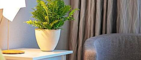 A hotel room with a bed, a bedside table with a lamp, a potted plant, and a framed picture on the wall.