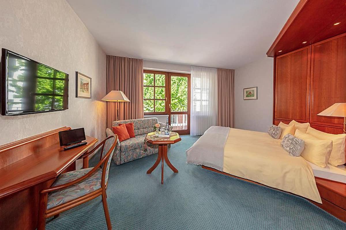 A hotel room with a bed, a desk, a chair, a small table, a TV, and a window with curtains.