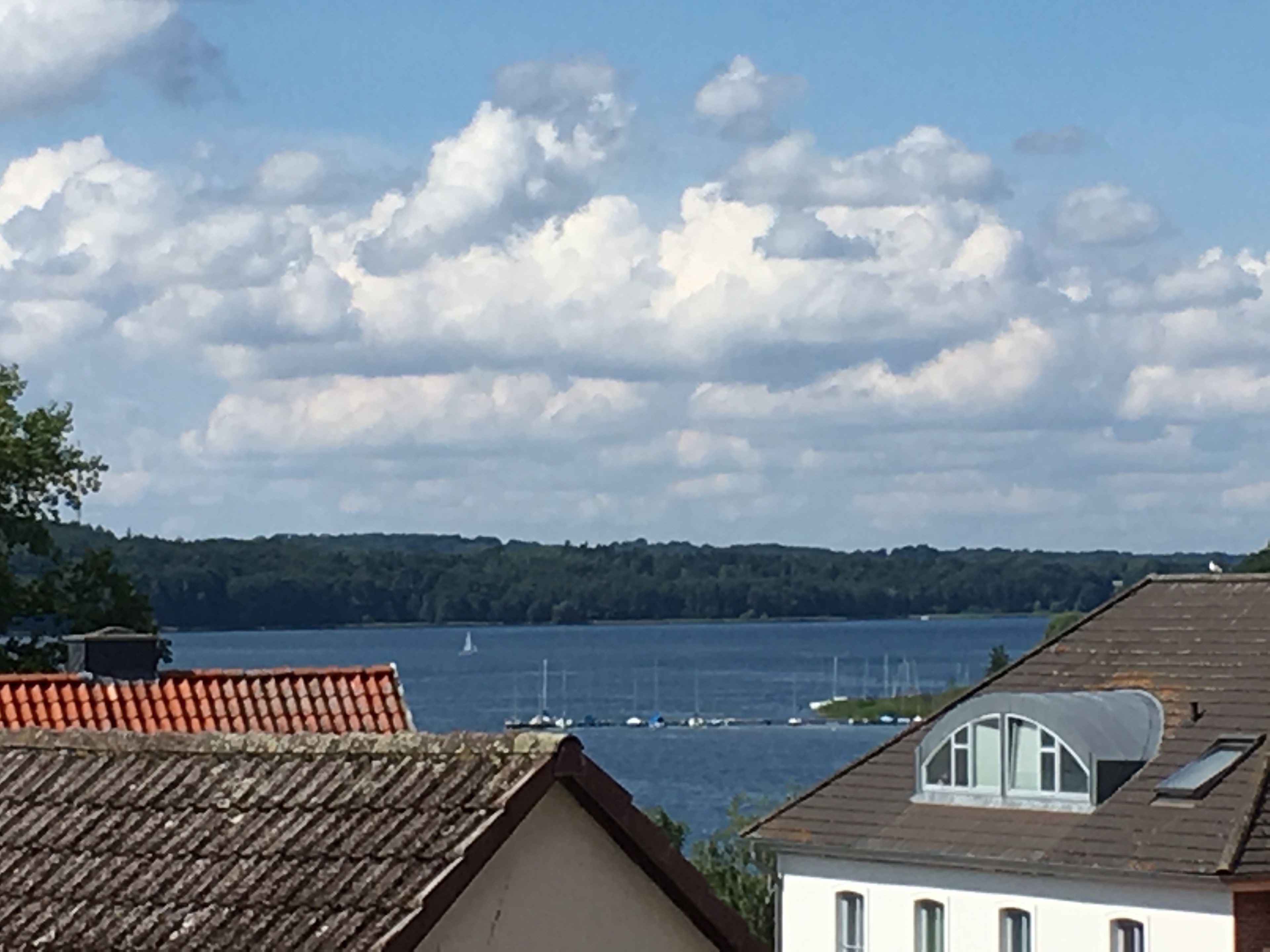 A view of a lake with a sailboat, surrounded by buildings with red-tiled roofs and a white building with a dormer window.