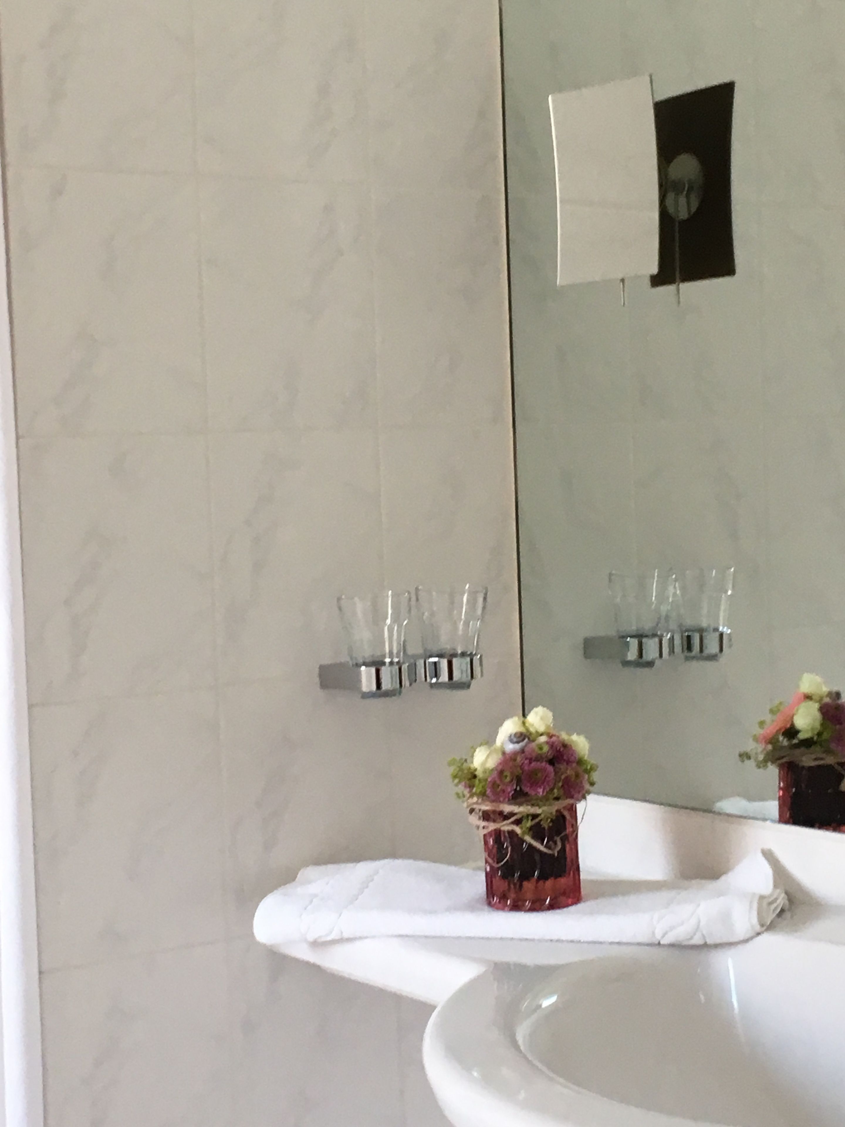 Un baño con pared de mármol, un lavabo y un espejo. Hay dos sujetavasos en la pared, uno con un florero.