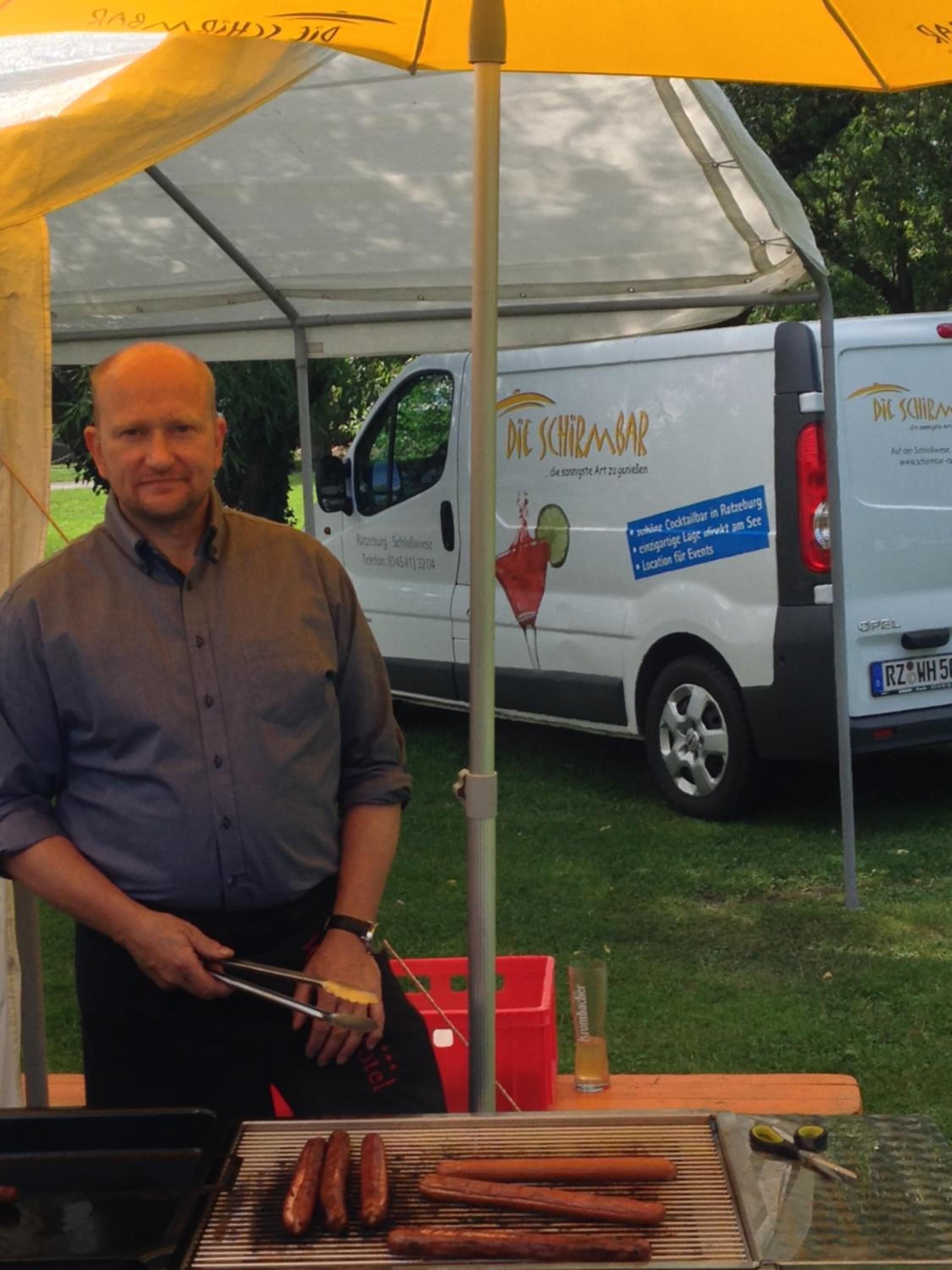 Mies grillaa makkaroita teltan alla, ja taustalla näkyy pakettiauto, jonka kylkeen on maalattu teksti ”Die Schirmbar”.