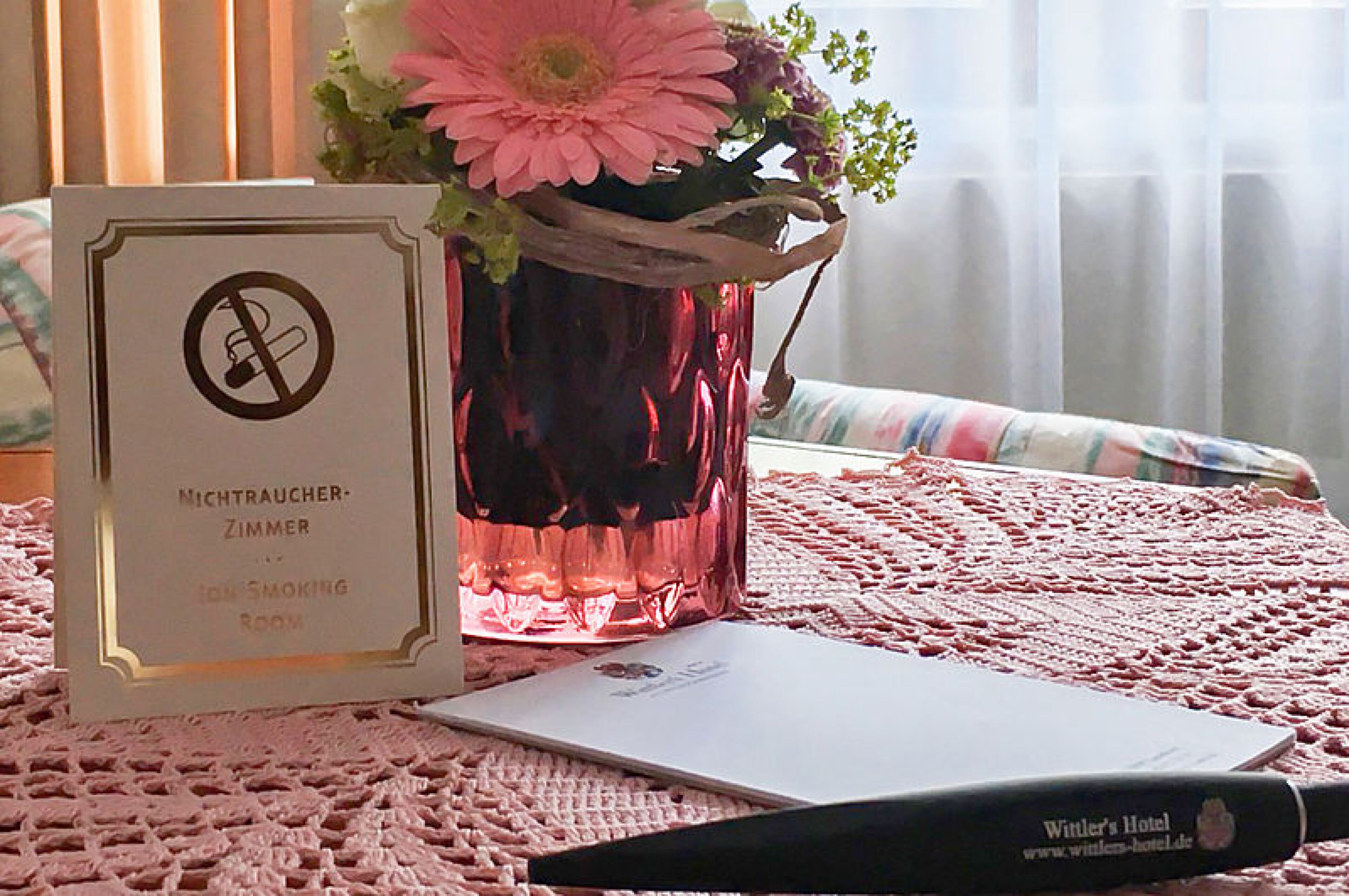 Hotelkamer met een ‘Niet roken’-bord, een roze vaas met bloemen, een laptop en een pen op een roze tafelkleed met textuur.