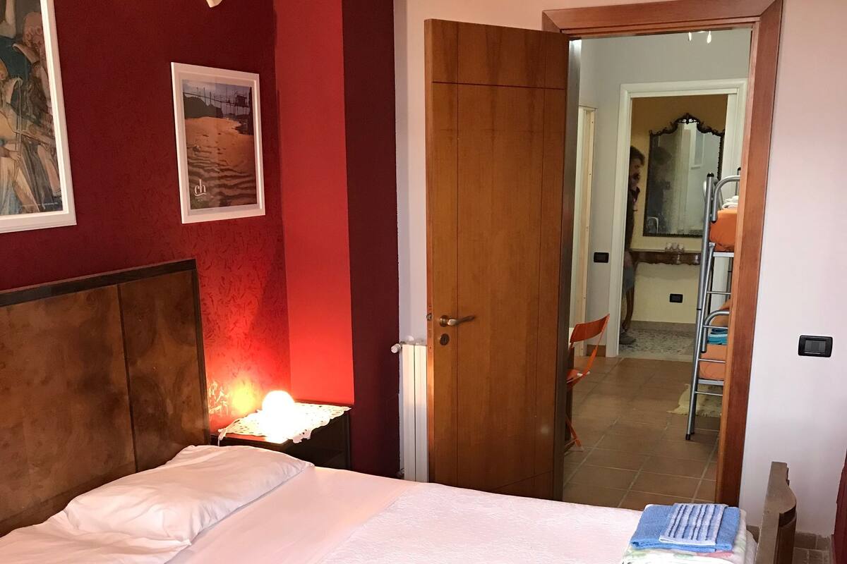 Een netjes opgemaakt bed met witte lakens, een houten hoofdbord en een bedlampje. Er is een deur die naar een andere kamer leidt, en een klein tafeltje met een lamp links van het bed.