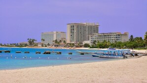 A beach with a large hotel, clear blue water, and a boat docked near the shore. - Sandy Shores at Sandcastles (Ocho Rios)