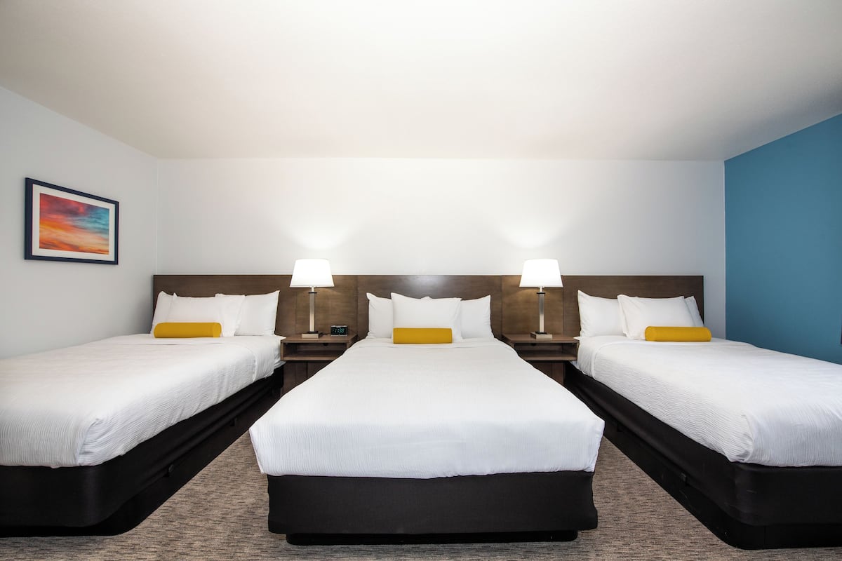 Ein Hotelzimmer mit drei Betten, jeweils mit weißer Bettwäsche und einem gelben Zierkissen. An den Nachttischen stehen Lampen und ein gerahmtes Bild hängt an der Wand.