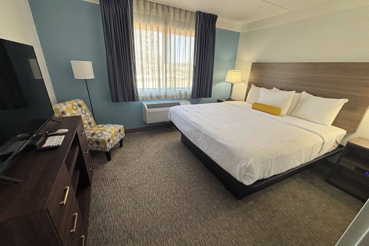 Ein Hotelzimmer mit einem Bett, einem Schreibtisch mit Fernseher, einem Sessel und einem Fenster mit Vorhängen.