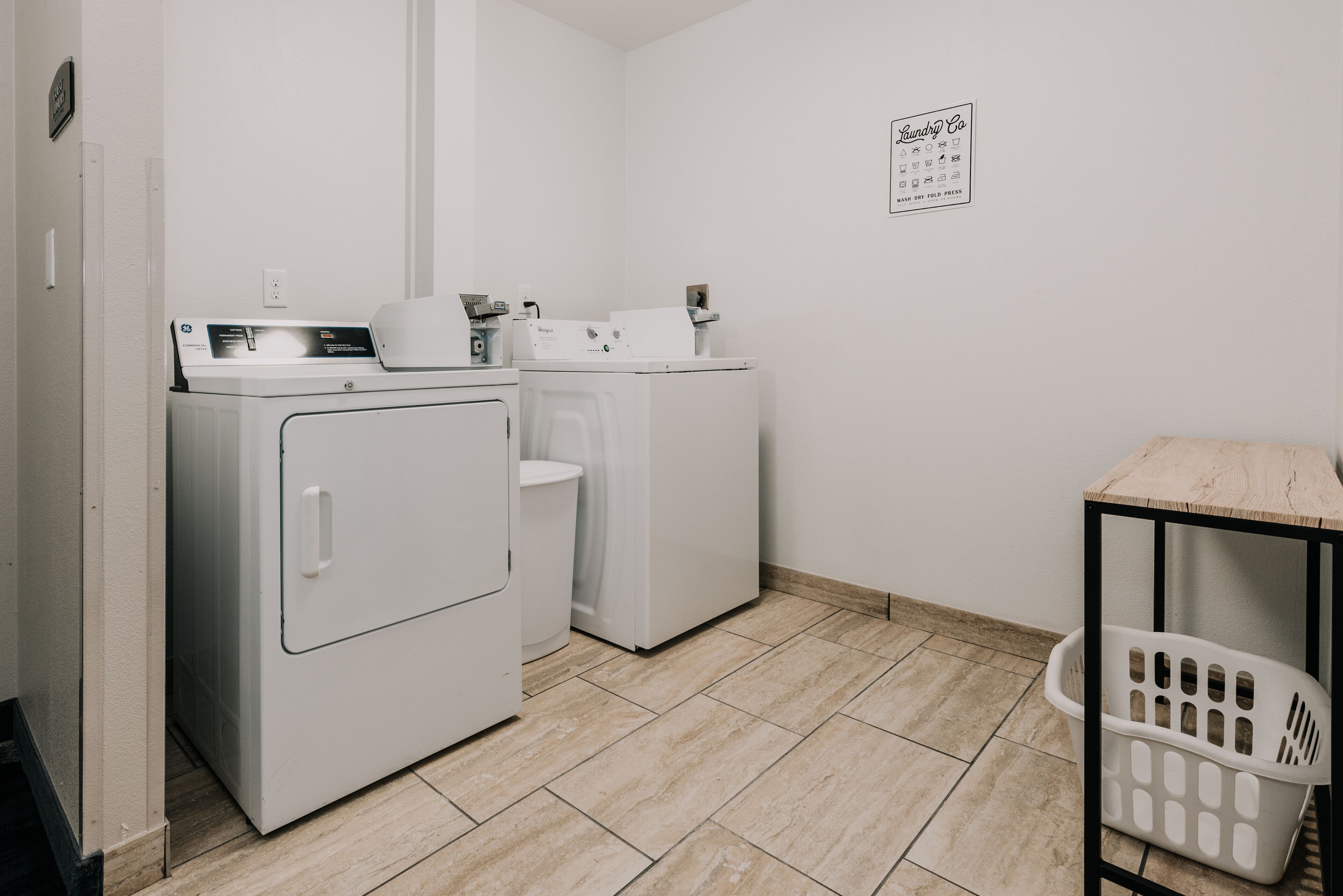 A laundry room with two white washing machines, a wooden table, and a laundry basket.