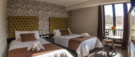 A hotel room with two beds, a stone wall, a balcony with a table and chairs, and a view of buildings outside.