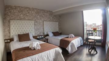 A hotel room with two beds, a stone wall, a balcony with a table and chair, and a view of buildings.
