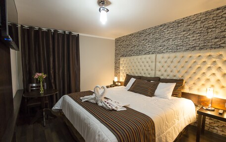 A hotel room with a bed, a small table with a vase of flowers, a chair, a nightstand with a lamp, and a stone-textured wall behind the bed.
