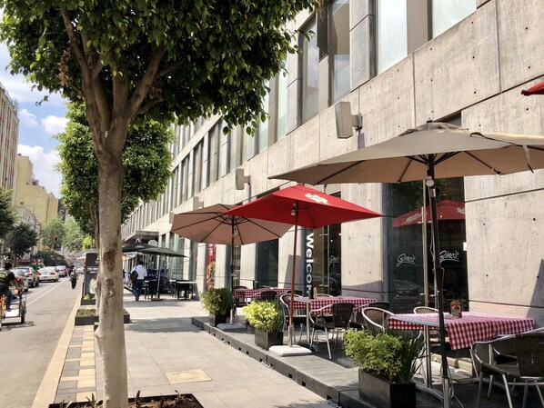 An outdoor cafe with tables and chairs, a tree, and a building with large windows.