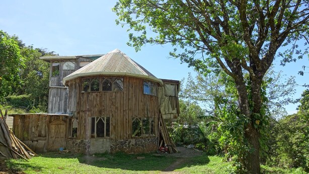A rustic wooden house with a steeply pitched roof and multiple windows, surrounded by lush greenery and a dirt path. - Casa Alquimia Artes (Monteverde)