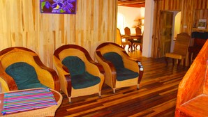 A room with wooden walls and floors, featuring two wicker chairs with green cushions, a striped throw blanket, and a framed picture of flowers. - Casa Alquimia Artes (Monteverde)