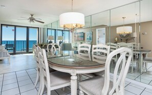 A dining area with a round glass table surrounded by white chairs, a ceiling fan, and a large window offering a view of the ocean.