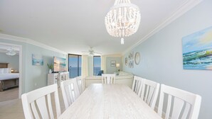 A dining area with a white wooden table and chairs, a chandelier, and a view of the ocean through large windows.