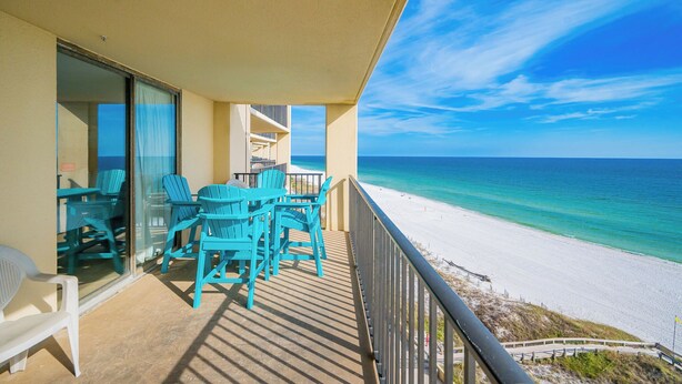 A balcony with turquoise chairs and a table, overlooking a beach with clear turquoise water and white sand.