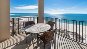 A balcony with a round wooden table and wicker chairs, overlooking a beach with clear blue water and a sandy shore.