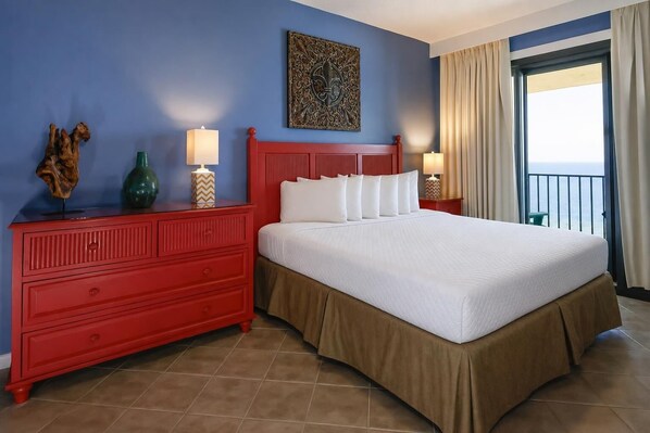A hotel room with a large bed, a red dresser, two bedside lamps, a decorative wall piece, and a view of the sea through a balcony.