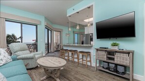 A modern living room with a sofa, a round coffee table, and a TV mounted on the wall. There is a kitchen area with a counter and stools. - Vacations are BETTER on the BEACH! ** 1BR @ Phoenix VII (Orange Beach)