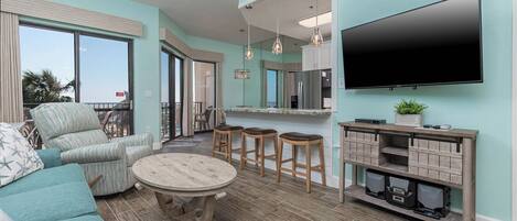 A modern living room with a sofa, a round coffee table, and a TV mounted on the wall. There is a kitchen area with a counter and stools.