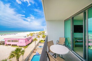 A balcony with a view of a beach, a pool, and a pink building.