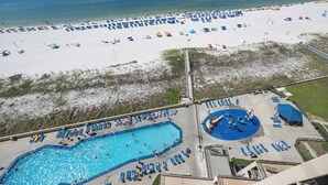 Un complejo turístico frente al mar con una amplia zona de piscina, numerosas tumbonas y una playa de arena con sombrillas.