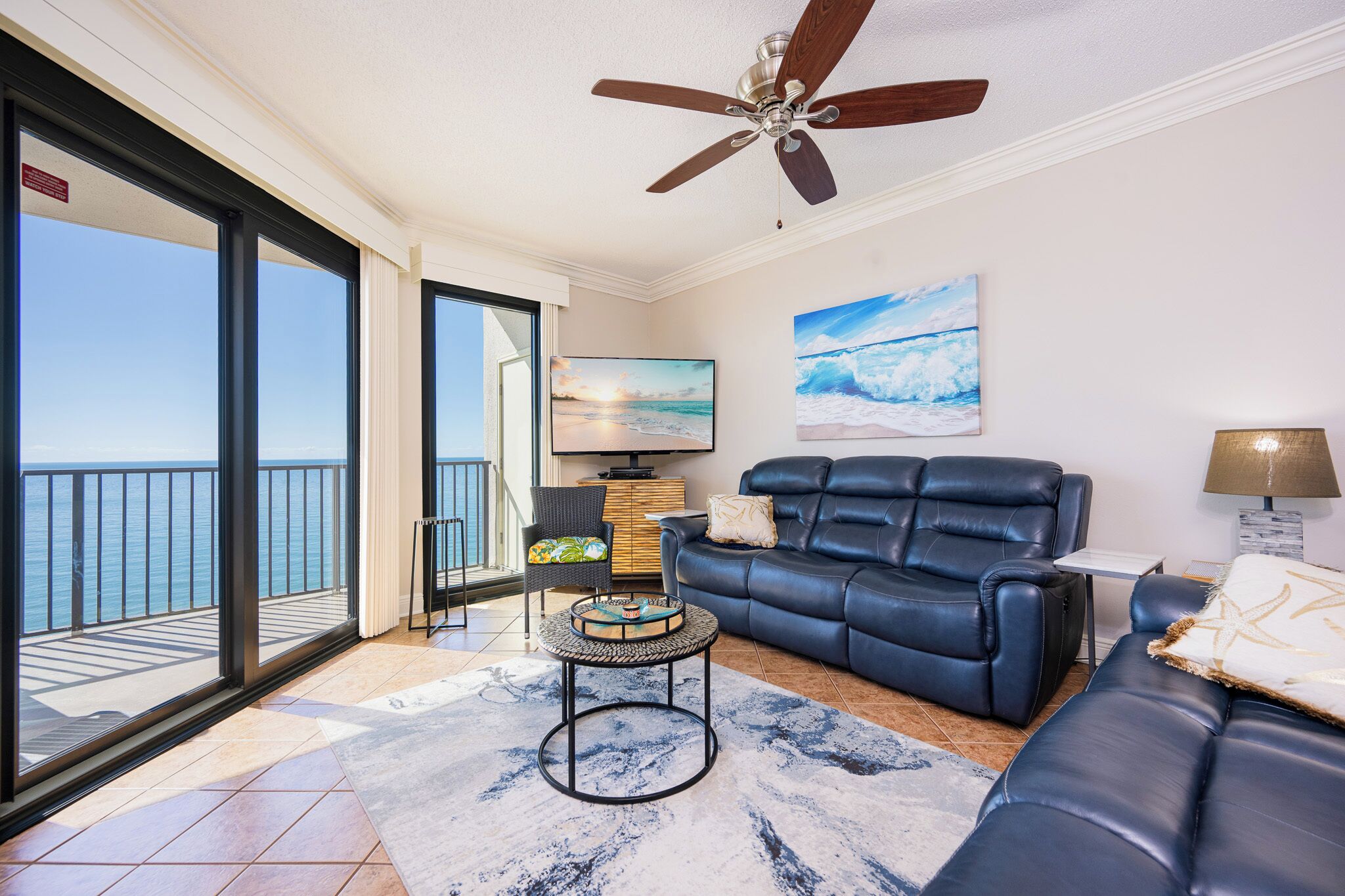 A living room with a sofa, a coffee table, and a television. There is a balcony with a view of the ocean.