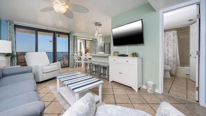 A spacious living room with a grey sofa, a striped ottoman, and a ceiling fan. A dining area with a table and chairs is visible through a glass door. A TV is mounted on the wall, and a bathroom with a toilet and shower is seen through an open door.