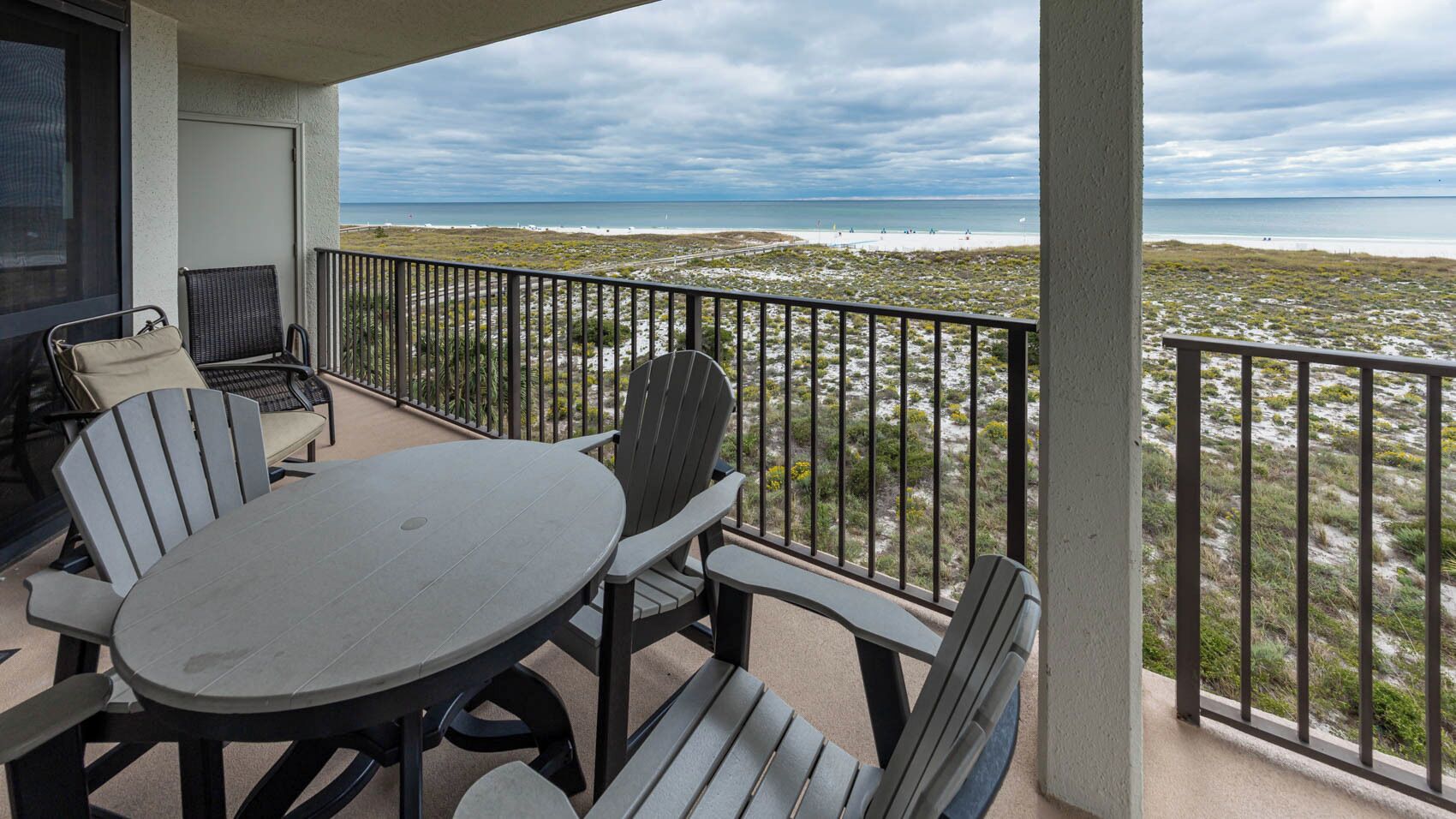 Un balcon avec une table ronde et quatre chaises Adirondack, donnant sur une plage à la végétation clairsemée et sous un ciel nuageux.