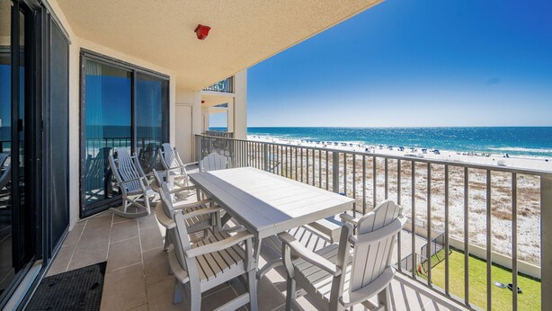 Ein Balkon mit weißem Tisch und Stühlen, der Blick auf einen Strand mit Menschen und einem klaren Himmel bietet.