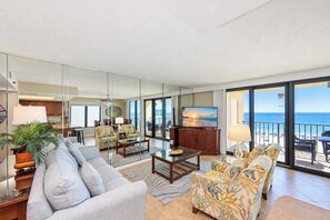 A spacious living room with a large flat-screen TV, comfortable seating, and a view of the ocean through large sliding doors.