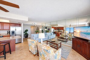 A modern living room with a sofa, coffee table, and chairs. There is a kitchen area with a refrigerator and microwave.