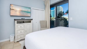 A bedroom with a large bed, a dresser, a television, and a view of buildings through a glass door.