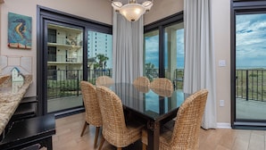 Un coin repas avec une table en verre et des chaises en osier, un tableau représentant un hippocampe, et une vue sur un bâtiment et la verdure, le tout visible à travers de grandes fenêtres.