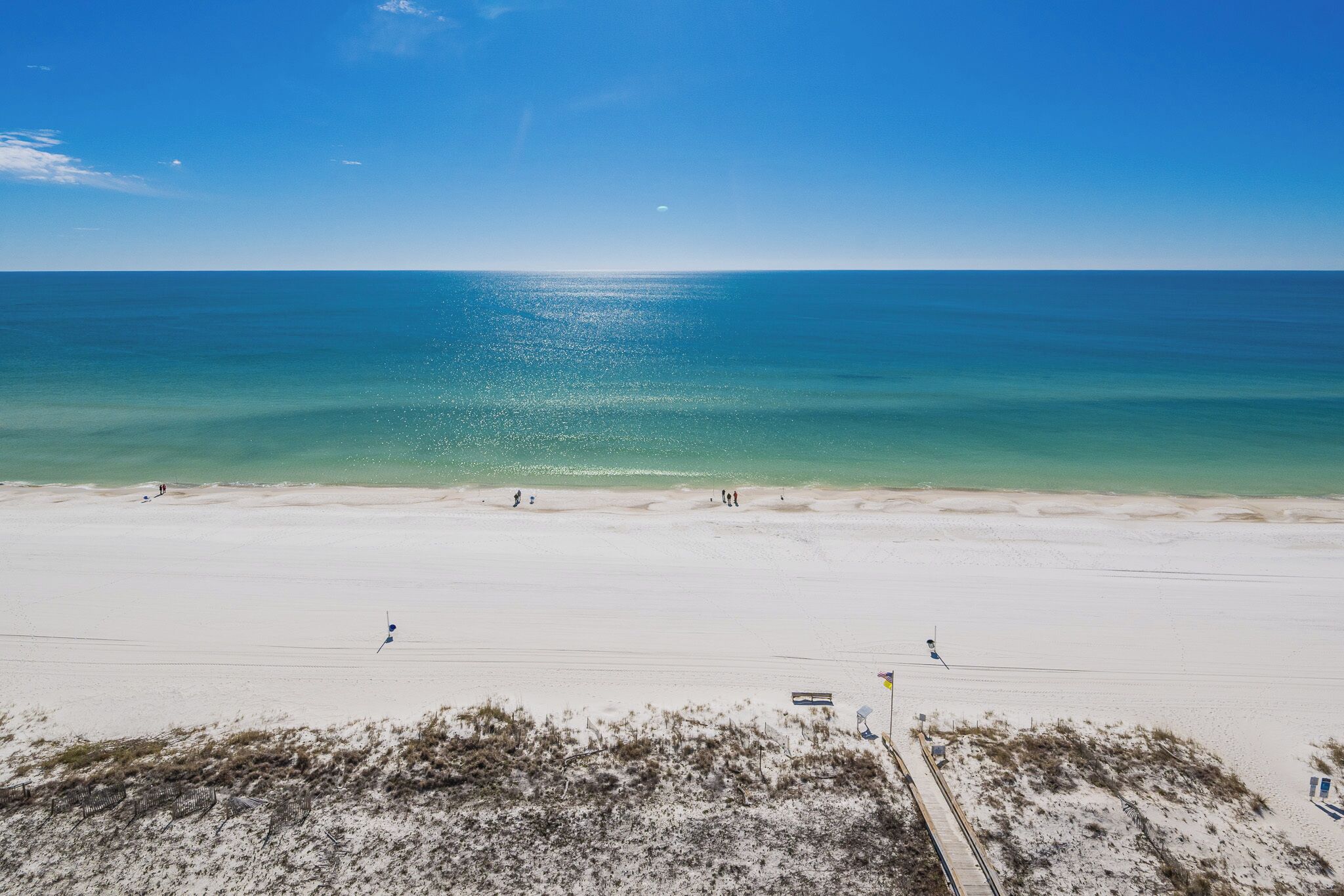 Een strand met een heldere lucht, kalme zee en enkele mensen die langs de kust wandelen.